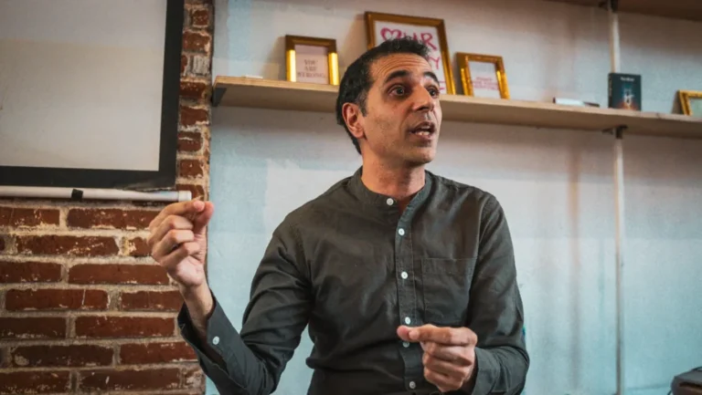 A man in a black shirt sits at a table, looking thoughtfully at a notebook in front of him.
