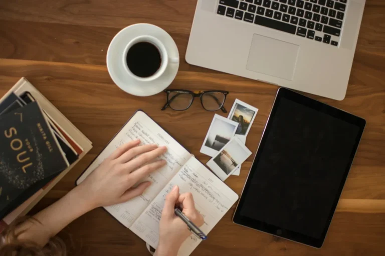 A woman's hands writing in a notebook beside a cup of coffee and a laptop on a wooden table.